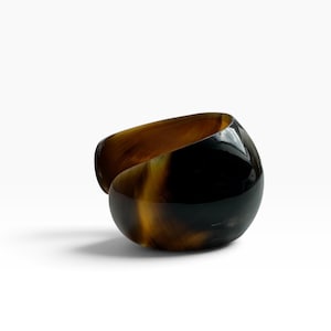 Op de afbeelding: Een brede, open armband met een gepolijste, glanzende afwerking en een gemarmerd patroon in diepzwart, donkerbruin en doorschijnende amberkleurige tinten. De gebogen, organische vorm creëert een glad, doorlopend silhouet met een brede opening.