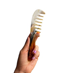 May include: A brown and black comb made of horn with a wide tooth design. The comb is held in a hand with purple nail polish.