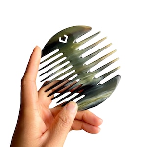 May include: A black and grey horn comb with a rounded shape and a small, white, diamond-shaped logo. The comb has a row of teeth on one side.