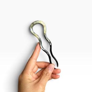 Pode incluir: Um gancho de cabelo em forma de U com uma superfície lisa e polida, apresentando um padrão marmoreado em tons translúcidos de creme, bege e preto profundo. O acessório possui dois dentes finos e curvos que terminam em pontas arredondadas.