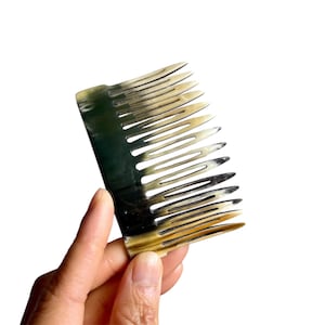 May include: A black and brown horn comb with a wide, curved handle and a row of teeth. The comb is held in a hand.