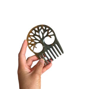 Könnte beinhalten: Ein Haarkamm aus poliertem Horn mit einem natürlichen, variierenden braunen und durchscheinenden Muster. Der obere Teil zeigt ein filigranes, ausgeschnittenes Design eines Baumes mit ausladenden Ästen und Wurzeln, unten mit sechs Zinken.