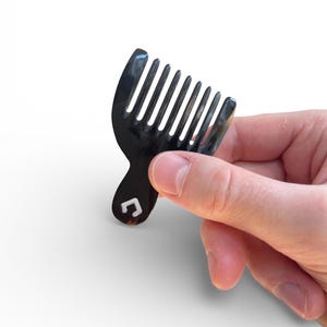 May include: A small, black comb with a curved handle and multiple teeth. The comb has a small, white square logo on the handle. The comb is held in a person's hand against a white background.