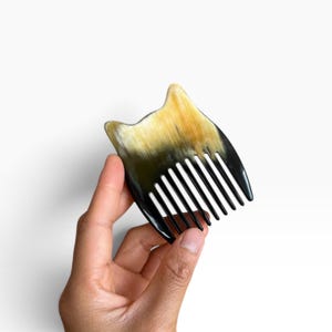 Könnte beinhalten: Ein kleiner Haarkamm aus natürlichem Horn mit einer markanten Katzenohr-Silhouette an der Oberseite. Das Material zeigt einen Farbverlauf von durchscheinenden Bernstein- und Goldtönen, die in ein tiefes, sattes Schwarz übergehen.