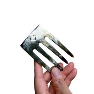 Puede incluir: Un peine para el cabello de cuerno negro y blanco con tres púas. El peine tiene un pequeño diseño cuadrado en la parte superior.