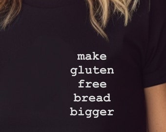 May include: A black short-sleeved t-shirt featuring white text printed on the chest that reads: make gluten free bread bigger. The garment has a crew neckline and a standard fit, with the text arranged in four stacked lines on the left side.