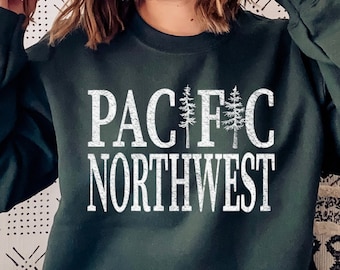 May include: Dark green sweatshirt with the text "PACIFIC NORTHWEST" in white, with tree graphics. The sweatshirt is a crew neck style. The model is wearing ripped blue jeans.