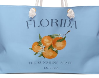 May include: A light blue tote bag with white rope handles. The front features a graphic of three oranges with green leaves and white blossoms. Text reads: FLORIDA, THE SUNSHINE STATE, EST. 1845. The design is centered on the bag's exterior surface.