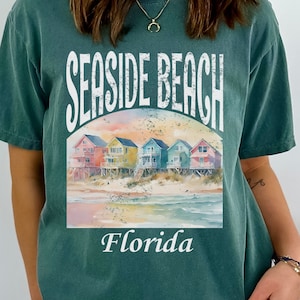 May include: A teal blue t-shirt with a vintage-style graphic of colourful beach houses and the text "Seaside Beach Florida".