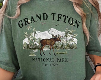 May include: An olive green short-sleeve t-shirt featuring a graphic print of a moose standing in front of mountains and trees. The text reads GRAND TETON NATIONAL PARK Est. 1929 in black lettering. The shirt has a crew neckline and a relaxed fit.