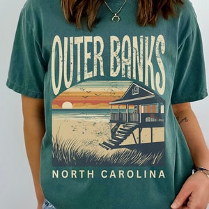 May include: A dark teal short-sleeve t-shirt featuring a graphic print with the text OUTER BANKS and NORTH CAROLINA. The design depicts a beach house on stilts, sand dunes, and a sunset over the ocean in shades of orange, blue, and cream.