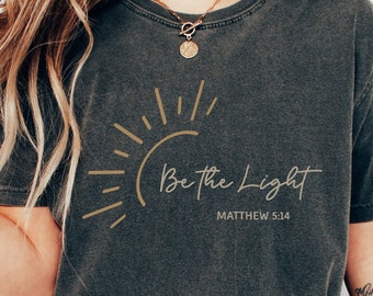 May include: A charcoal gray short-sleeve t-shirt featuring a gold graphic of a sunburst and the text "Be the Light" followed by "MATTHEW 5:14" in a clean, sans-serif font. The shirt has a vintage-washed appearance with a crew neckline.