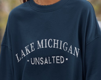 Op de afbeelding: Een donkerblauw sweatshirt met ronde hals en witte letters in een verweerde stijl op de borst. De tekst LAKE MICHIGAN staat in een boogvorm, met het woord UNSALTED eronder gecentreerd tussen twee kleine horizontale streepjes.