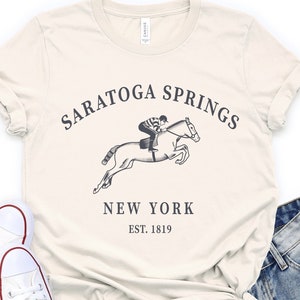 May include: A cream-colored short-sleeve t-shirt featuring a black graphic of a jockey riding a jumping horse with the text SARATOGA SPRINGS NEW YORK EST. 1819, paired with white canvas sneakers and distressed blue denim shorts.