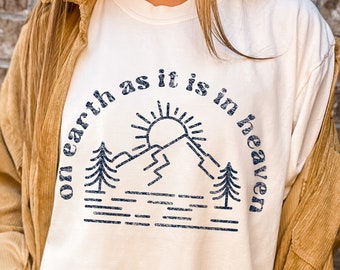May include: A cream-colored t-shirt featuring a dark blue graphic print of a mountain range, a sun, and pine trees. The text "on earth as it is in heaven" is printed in a circular arc above the mountain graphic in a distressed navy blue font.