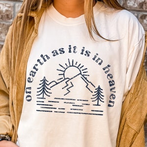 Könnte beinhalten: Ein weißes T-Shirt mit einem blauen Grafikdesign, auf dem "on earth as it is in heaven" steht. Die Grafik zeigt eine Sonne, Berge und Bäume.
