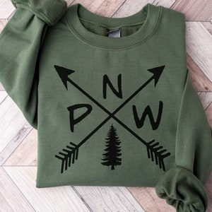 May include: An olive green crewneck sweatshirt featuring a black graphic print of two crossed arrows. The letters P, N, and W are positioned between the arrows, and a black silhouette of a pine tree is centered below the intersection.