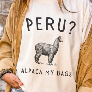 Peut inclure: Un t-shirt blanc cassé avec une illustration noire d'un alpaga au centre. Au-dessus de l'alpaga, le texte indique PERU? et en dessous, le texte indique ALPACA MY BAGS. Le t-shirt est porté sous une chemise en velours côtelé beige et un short en jean.