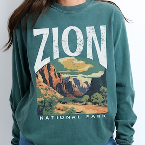 May include: A teal long-sleeve t-shirt with a vintage-style graphic of Zion National Park. The graphic features a mountain range, a road, and trees. The text "ZION NATIONAL PARK" is printed below the graphic.