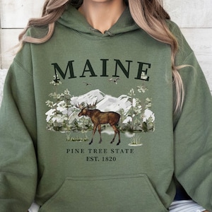 Puede incluir: Sudadera con capucha verde oliva con un diseño gráfico frontal. El diseño incluye la palabra "MAINE", un alce, montañas y árboles. Debajo está el texto "PINE TREE STATE EST. 1820". La sudadera tiene un bolsillo de canguro y una capucha con cordón.