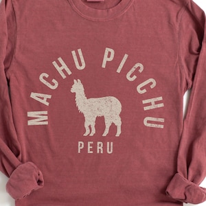 Op de afbeelding: Een t-shirt met lange mouwen en een ronde hals in een gedempte baksteenrode kleur. De voorkant heeft een verweerde beige afbeelding van een lama, gecentreerd onder de gebogen tekst "MACHU PICCHU" en het woord "PERU" in een schreefloos lettertype.