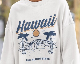 May include: White sweatshirt with a blue and brown graphic design of palm trees, a wave, and a sun. The text "Hawaii" is written in a blue cursive font above the graphic. The text "The Aloha State" is written below the graphic.