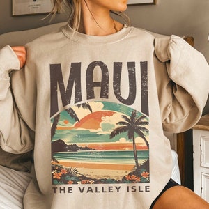 May include: A beige crewneck sweatshirt featuring a graphic print with the text MAUI THE VALLEY ISLE. The design depicts a tropical beach scene with a sunset, palm trees, and flowers in shades of orange, teal, and brown with a distressed texture.