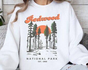 May include: White sweatshirt with a retro graphic design featuring a sunset over a redwood forest. The text "Redwood National Park Est. 1968" is printed below the graphic.