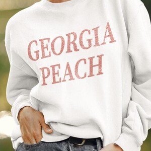 May include: A white long-sleeve crewneck sweatshirt featuring the text GEORGIA PEACH printed in a distressed, muted pink font across the chest. The garment has ribbed cuffs and a ribbed neckline, providing a classic fit and a clean, simple aesthetic.