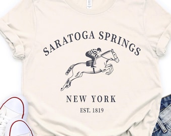 Puede incluir: Una camiseta de manga corta color crema con un gráfico gris oscuro de un jinete sobre un caballo saltando. El texto SARATOGA SPRINGS, NEW YORK y EST. 1819 aparece en una fuente serif gris oscuro centrada en el pecho de la prenda.
