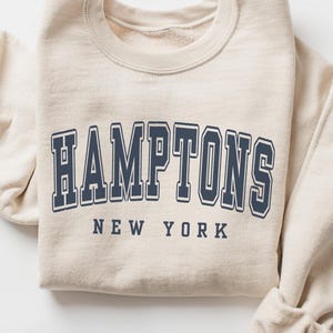 Puede incluir: Una sudadera de cuello redondo en color beige con el texto HAMPTONS en una fuente grande de estilo universitario azul marino arqueada sobre el pecho, con las palabras NEW YORK impresas en una fuente recta azul marino más pequeña debajo.