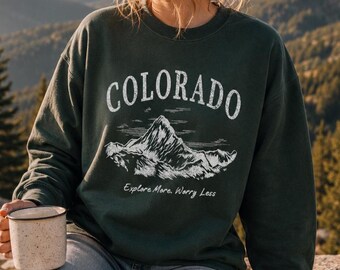 May include: A dark green crewneck sweatshirt featuring a white graphic of a mountain range. The text "COLORADO" is printed in a large, arched font above the mountain, with the phrase "Explore More, Worry Less" written in a smaller script below.