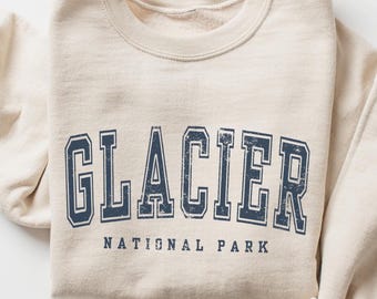 Könnte beinhalten: Ein beigefarbenes Sweatshirt mit Rundhalsausschnitt und dem Schriftzug GLACIER NATIONAL PARK in einer dunkelblauen, abgenutzten Schriftart im College-Stil. Das Kleidungsstück hat lange Ärmel und einen gerippten Ausschnitt mit schlichtem Design.