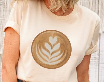 Puede incluir: Una camiseta de manga corta color crema con un gráfico circular en el pecho. El gráfico muestra una vista cenital de un café latte con arte de espuma blanca en forma de corazón y hojas sobre un fondo de café de color marrón.