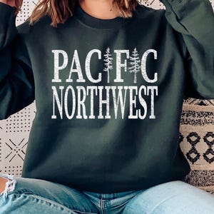 May include: A dark forest green long-sleeve crewneck sweatshirt featuring white distressed text that reads PACIFIC NORTHWEST. The letter I in PACIFIC is replaced by two vertical pine tree illustrations, creating a nature-inspired graphic design.