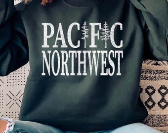 May include: A dark forest green long-sleeve crewneck sweatshirt featuring white distressed text that reads PACIFIC NORTHWEST. The letter I in PACIFIC is replaced by two vertical pine tree illustrations, creating a nature-inspired graphic design.