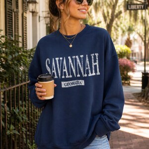 May include: Navy blue sweatshirt with "SAVANNAH GEORGIA" printed in white. The sweatshirt is worn with denim shorts and sunglasses. A person is holding a coffee cup.