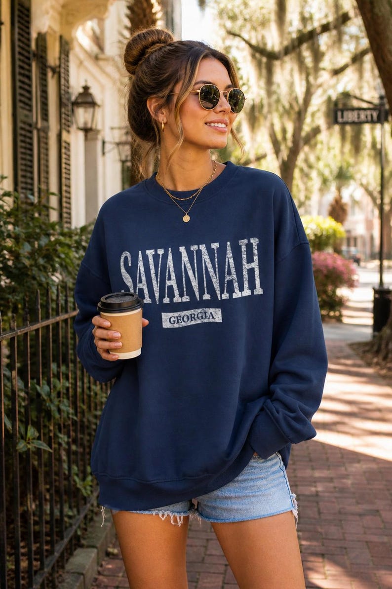 May include: Navy blue sweatshirt with "SAVANNAH GEORGIA" printed in white. The sweatshirt is worn with denim shorts and sunglasses. A person is holding a coffee cup.