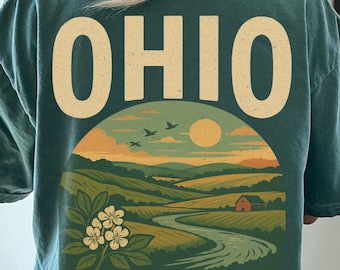 Könnte beinhalten: Ein dunkelgrünes T-Shirt mit einem Grafikdruck auf der Rückseite, der oben den Text OHIO und unten BUCKEYE STATE zeigt. Das Motiv stellt eine Landschaft mit sanften Hügeln, einem gewundenen Fluss, einer kleinen roten Scheune, Vögeln und Blumen dar.
