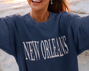 May include: A navy blue crewneck sweatshirt featuring the text NEW ORLEANS printed in a distressed white font across the chest. The garment has long sleeves, a ribbed collar, and a relaxed fit, showcasing a vintage-inspired graphic design.