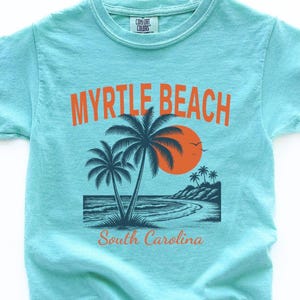 May include: A light turquoise short-sleeve t-shirt featuring a graphic of a beach scene with palm trees, an orange sun, and the text MYRTLE BEACH and South Carolina. The shirt has a Comfort Colors brand label printed on the inside of the collar.