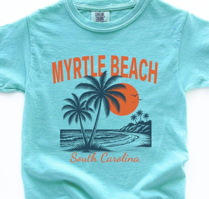 May include: A light turquoise short-sleeve t-shirt featuring a graphic of a beach scene with palm trees, an orange sun, and the text MYRTLE BEACH and South Carolina. The shirt has a Comfort Colors brand label printed on the inside of the collar.