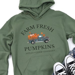 May include: Green hooded sweatshirt with a graphic of a blue truck carrying pumpkins and fall leaves. The text reads "FARM FRESH PUMPKINS" with "APPLES • CIDER • HAYRIDES" below. The sweatshirt has a front pocket and drawstring hood.