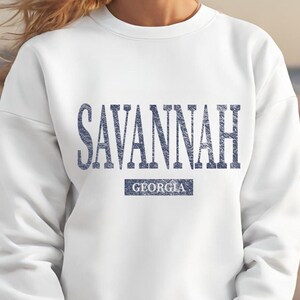 May include: White sweatshirt with the word "SAVANNAH" in blue, distressed lettering across the chest. Below the city name is the word "GEORGIA" in a smaller, rectangular blue box. The sweatshirt is a casual style.