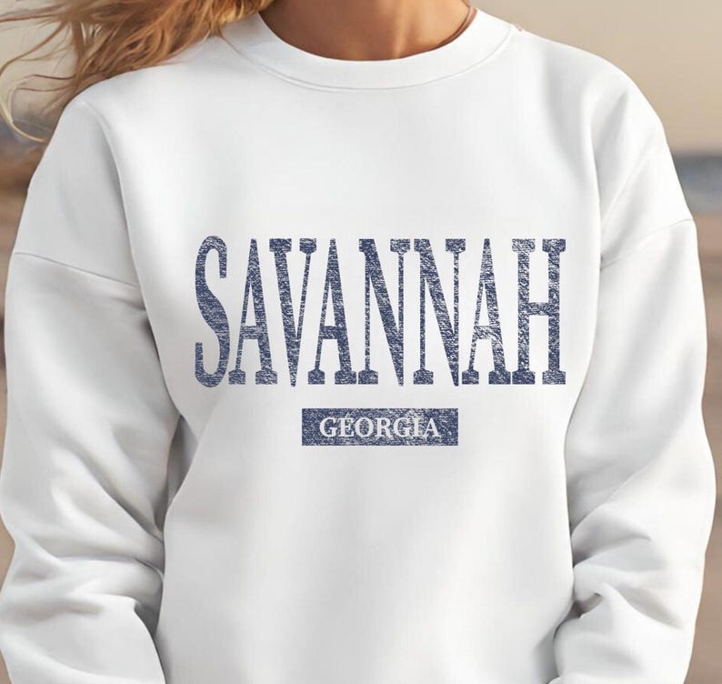 May include: White sweatshirt with the word "SAVANNAH" in blue, distressed lettering across the chest. Below the city name is the word "GEORGIA" in a smaller, rectangular blue box. The sweatshirt is a casual style.