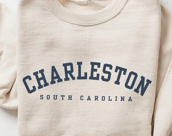 May include: A beige crewneck sweatshirt featuring the dark blue text CHARLESTON arched across the chest, with the words SOUTH CAROLINA printed in a smaller, straight font directly underneath the city name in a matching dark blue color.