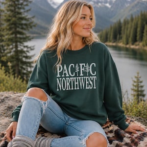 May include: A dark green crewneck sweatshirt featuring the white text PACIFIC NORTHWEST with two pine tree illustrations replacing the letter I, paired with light blue distressed denim jeans and grey knit leg warmers.