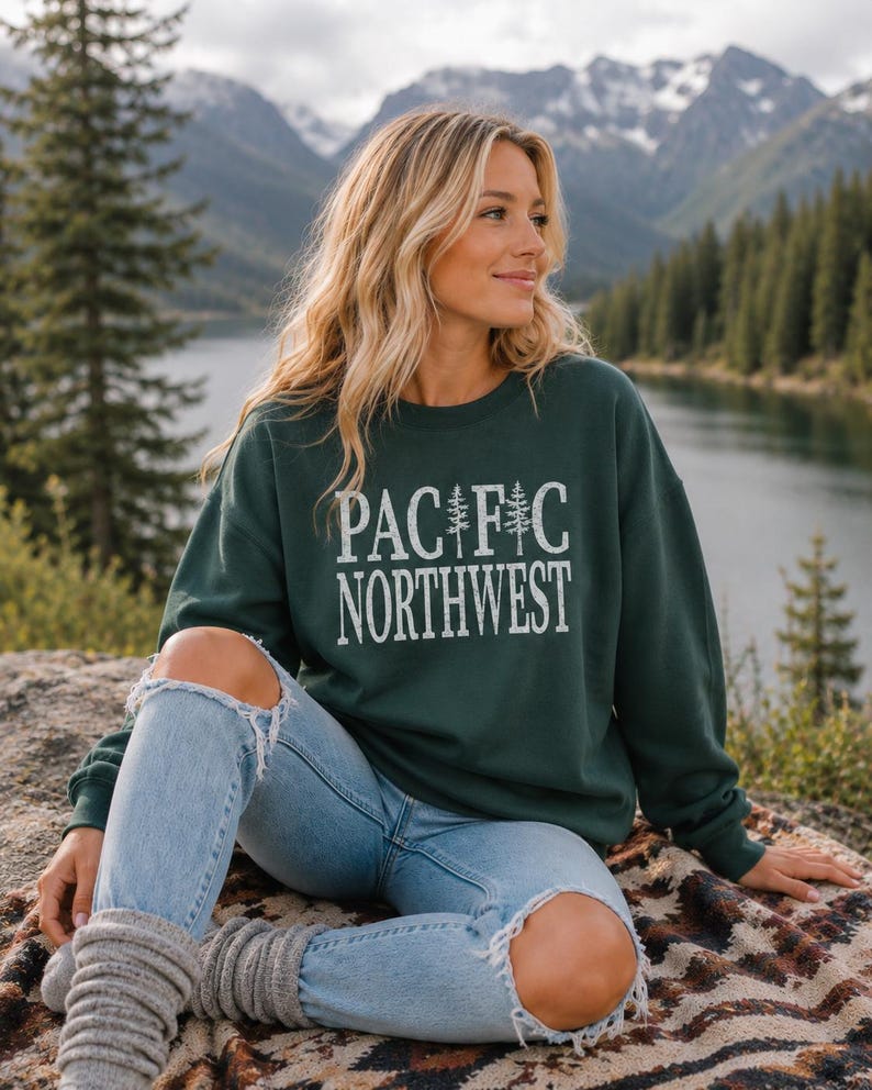 May include: A dark green crewneck sweatshirt featuring the white text PACIFIC NORTHWEST with two pine tree illustrations replacing the letter I, paired with light blue distressed denim jeans and grey knit leg warmers.