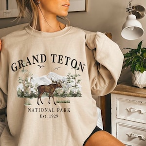 Puede incluir: Sudadera beige de cuello redondo con un gr&aacute;fico de un alce en una escena forestal con monta&ntilde;as y el texto "Grand Teton National Park Est. 1929". El dise&ntilde;o presenta &aacute;rboles, p&aacute;jaros y una cordillera.