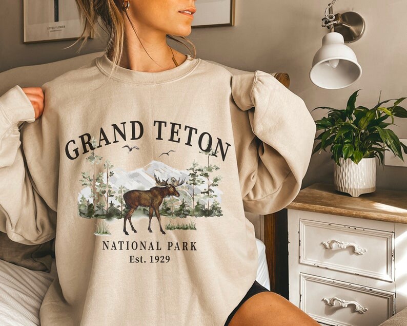 Puede incluir: Sudadera beige de cuello redondo con un gr&aacute;fico de un alce en una escena forestal con monta&ntilde;as y el texto "Grand Teton National Park Est. 1929". El dise&ntilde;o presenta &aacute;rboles, p&aacute;jaros y una cordillera.